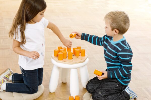 Für die Produktseite "Nino Shake´n Play 526": Ein Mädchen mit braunen Haaren und ein Junge mit blonden Haaren knien auf dem Boden und spielen mit einem Holzspielzeug, bestehend aus einem weißen Hocker, auf dem orangefarbene Zylinder in Löcher gesteckt werden.