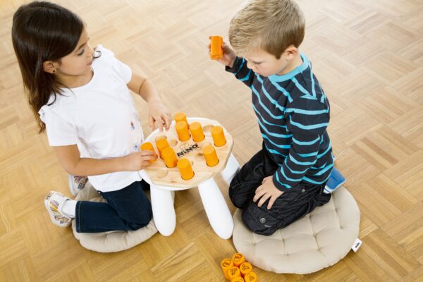 Zwei Kinder, ein Mädchen mit dunklen Haaren und ein Junge mit blondem Haar, knien auf hellen, runden Kissen vor einem Nino Shake´n Play 526 Spieltisch und spielen mit orangefarbenen Spielsteinen auf hellem Holzboden.