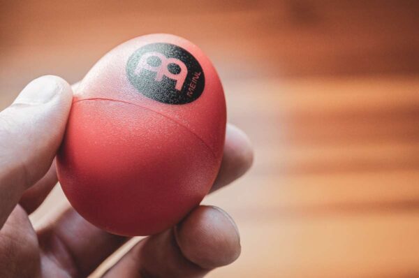Eine Hand hält ein rotes Meinl Shaking Egg mit einem schwarzen Logo auf einem unscharfen, hölzernen Hintergrund.