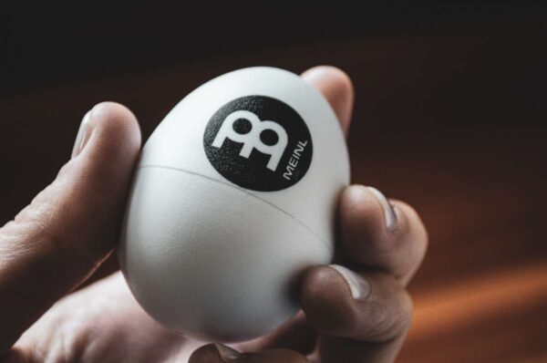 Eine Hand hält ein weißes Meinl Shaking Egg mit schwarzem Markenlogo vor einem unscharfen, dunklen Hintergrund.