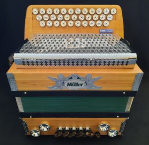 Eine gebrauchte, helle, dreireihige Harmonika der Marke Müller mit Knöpfen, einem silberfarbenem, gemustertem Diskantverdeck, grünen Balgen und silberfarbenem Markenemblem ist vor einem dunklen Hintergrund abgebildet.