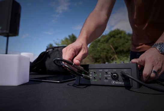 Ein Mann mit einem blauen Uhrenarmband schließt mehrere schwarze Audiokabel an ein schwarzes Audio-Interface auf einem schwarzen Tisch an, im Hintergrund sind ein Lautsprecher, ein weiß leuchtender Würfel, ein Tablet und Bäume unter einem blauen Himmel sichtbar.