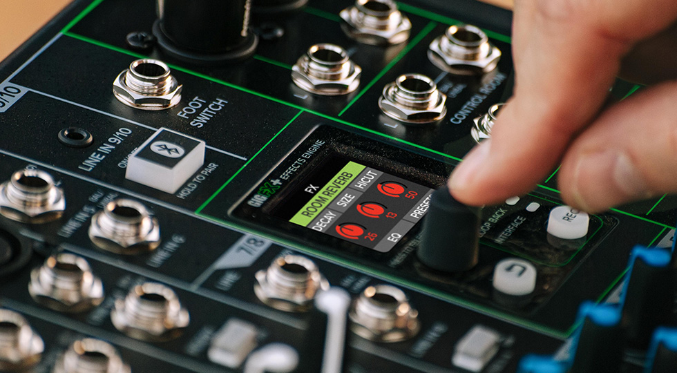 Eine Hand dreht an einem schwarzen Drehknopf eines detailreichen, schwarz-grünen Mischpults mit zahlreichen silberfarbenen Anschlüssen und einem kleinen Farbdisplay, das rote grafische Elemente zeigt.