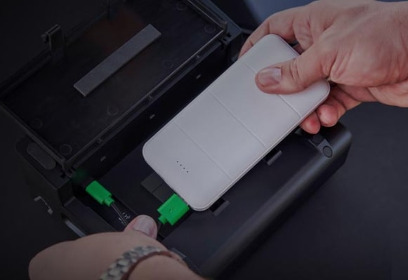 Eine Hand legt eine weiße, rechteckige Powerbank in ein schwarzes, offenes Gerät mit grünen Anschlüssen.