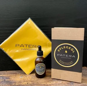 Das Bild zeigt ein Patera Handpan Pflegeöl-Set, bestehend aus einer braunen Glasflasche mit Pipette, einem eckigen Karton mit einem schwarzen Etikett und einem gelben Reinigungstuch, alle verziert mit dem "Patera Handpan"-Logo.