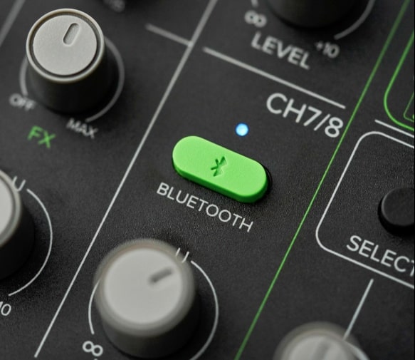 Nahaufnahme eines schwarzen Audiomischpults mit einem hellgrünen Bluetooth-Knopf mit einem Bluetooth-Symbol und dem Wort "BLUETOOTH" darunter, neben dem sich ein kleiner, leuchtend blauer Punkt befindet.