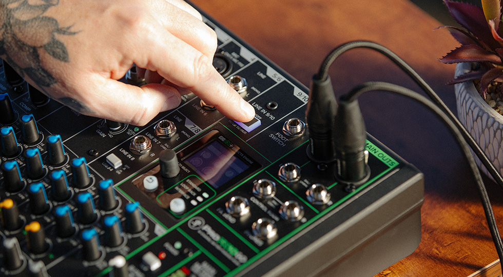 Eine tätowierte Hand drückt einen hellen Knopf auf einem schwarzen Mischpult mit grünen Akzenten, an das zwei XLR-Kabel angeschlossen sind.