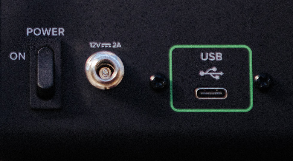 Auf einer schwarzen Oberfläche befinden sich ein schwarzer Netzschalter in der "ON"-Position, eine runde 12V-2A-Buchse und ein grünumrandeter USB-C-Anschluss, der von zwei schwarzen Schrauben flankiert wird.