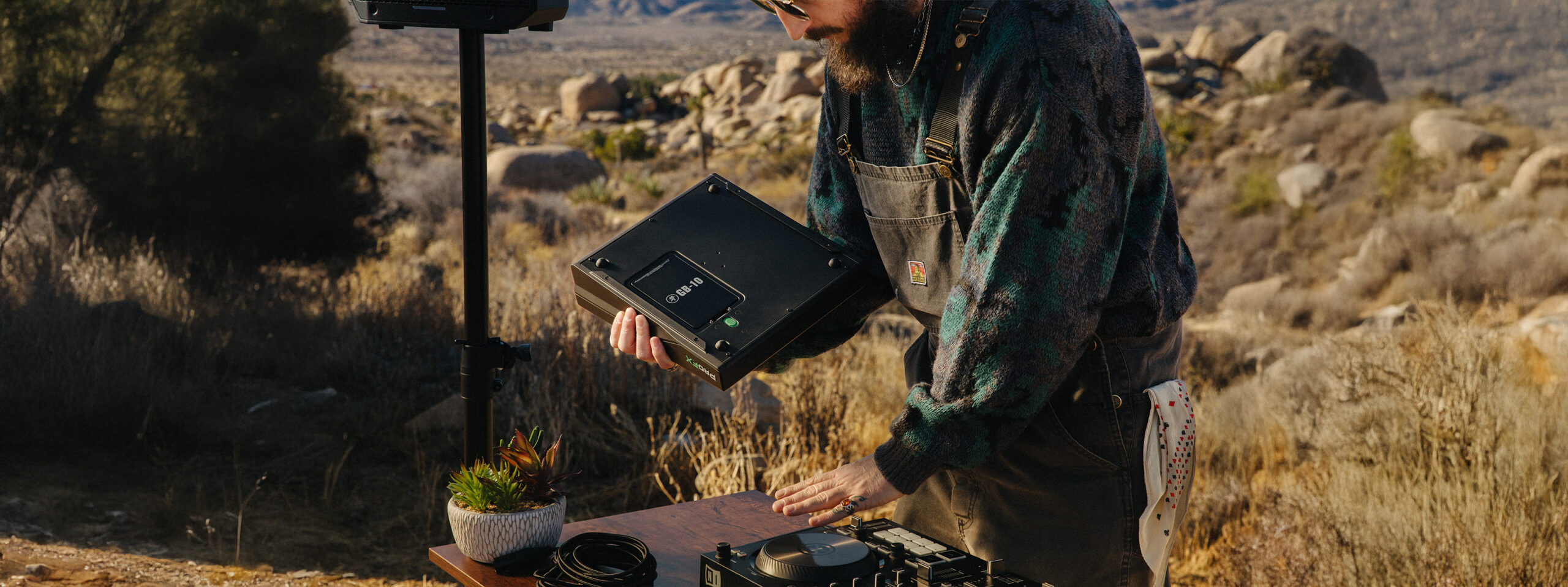 Ein bärtiger Mann mit Brille und grünkariertem Hemd hantiert in einer trockenen Landschaft an einem Tisch mit technischem Equipment und einem schwarzen Kasten in seinen Händen.