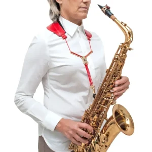 Eine Frau mit kurzen, grauen Haaren trägt ein weißes Hemd und einen roten BG S79YMSH Zen Yoke-Gurt aus Leder, während sie ein goldenes Saxophon hält.