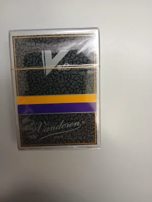 Eine durchsichtige, versiegelte Packung Vandoren B-Klarinette V12 Stärke 5 (10 Stk.) mit einem schwarz-weiß gemusterten Label, einem gelben und einem violetten Streifen.