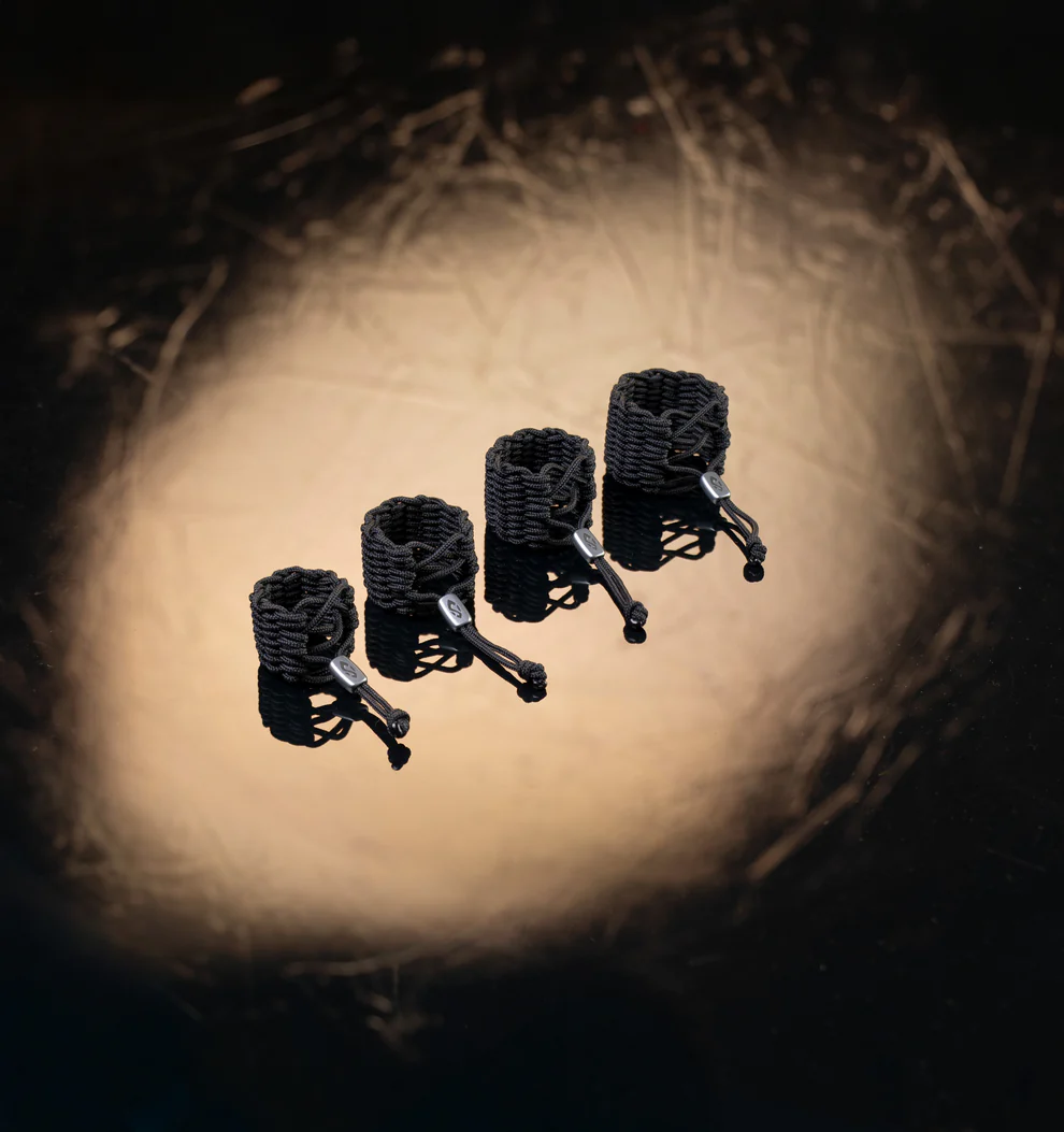 Vier schwarze FIBRA Ligaturen für Sopran Saxophon und Es Klarinette stehen in einer Reihe vor einem dunkelbraunen, verschwommenen Hintergrund.