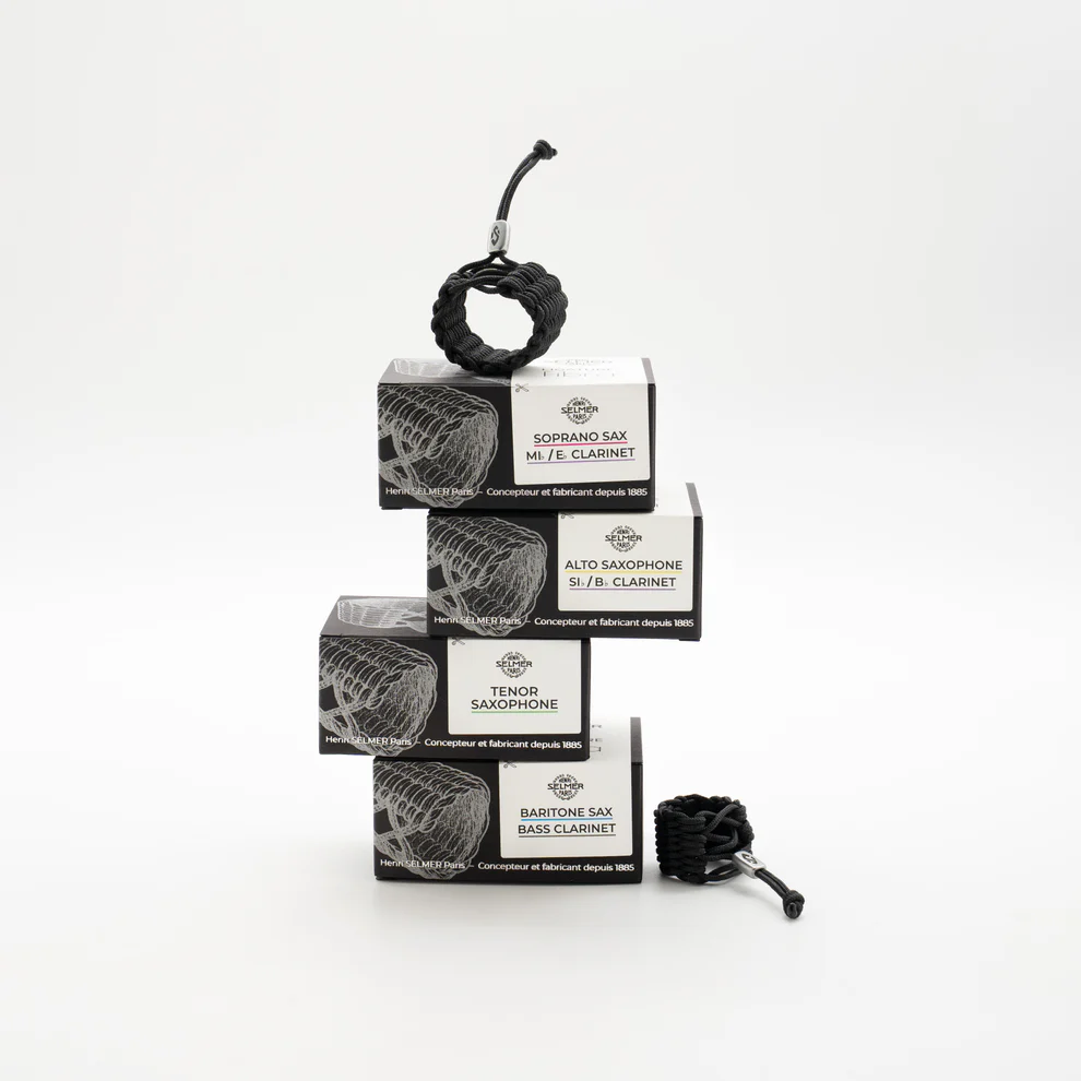 Auf einem weißen Hintergrund steht ein Turm aus vier schwarzen Produktverpackungen, auf denen der Text "Soprano Sax, Alto Clarinet", "Alto Sax, Bb Clarinet", "Tenor Saxophone" und "Baritone Sax, Bass Clarinet" steht, wobei oben auf dem Turm eine schwarze FIBRA Ligatur für Sopran Saxophon sitzt und rechts daneben eine weitere FIBRA Ligatur liegt.