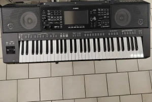 Das Bild zeigt ein gebrauchtes, schwarzes Yamaha PSR-SX920 Keyboard mit Lautsprechern, Tasten und einem zentralen Bedienfeld, das auf einem gefliesten Untergrund steht.