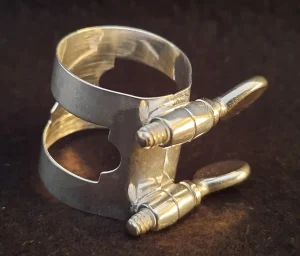 Eine silberfarbene Blattschraube für B-Klarinette mit zwei seitlichen Schrauben ist vor einem unscharfen, dunklen Hintergrund abgebildet.