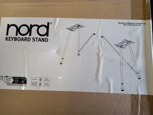 Auf einem Karton ist ein weißes Etikett mit dem schwarzen "nord KEYBOARD STAND" Logo und einer technischen Zeichnung des Clavia Nord Keyboard Ständer EX abgebildet.