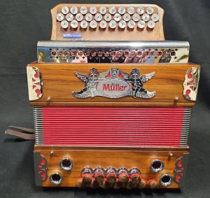 Nahaufnahme einer gebrauchten Müller Mini Steirischen Harmonika mit rot-braunen Balgen, einem silberfarbenen Müller-Logo zwischen zwei Engeln und Knöpfen aus Holz und Metall.