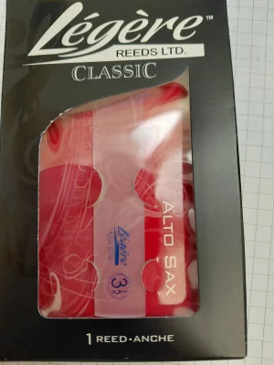 Ein Paket eines Legere Classic Alto Saxophon-Blattes der Stärke 3 1/2 mit roter Verpackung und dem Schriftzug "Legere REEDS LTD." auf schwarzem Grund ist vor einem karierten Hintergrund abgebildet.