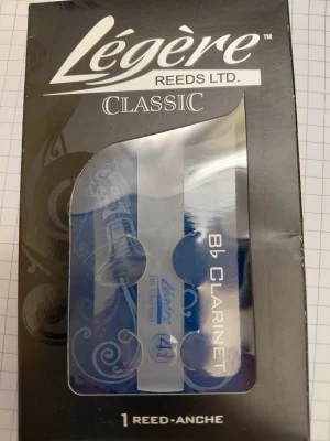 Ein schwarzer Karton mit der Aufschrift "Légère Reeds LTD. Classic" und der Aufschrift "Bb Clarinet" zeigt ein einzelnes, durchsichtiges Légère-Klarinettenblatt der Stärke 4 3/4 in einer durchsichtigen, bläulich schimmernden Verpackung, die auf einem karierten Untergrund liegt.