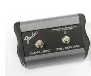 Ein grauer Fender Fußschalter mit schwarzem Gehäuse, zwei silberfarbenen Fußtastern, roten und silbernen LEDs sowie der Aufschrift "Channel Select" und "Drive / More Drive" ist auf weißem Grund zu sehen.