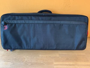 Eine dunkelgraue Backline XPND 2 Pedalboard Transporter-Tasche liegt horizontal auf einem hellen Holzboden vor einer ockerfarbenen Wand.